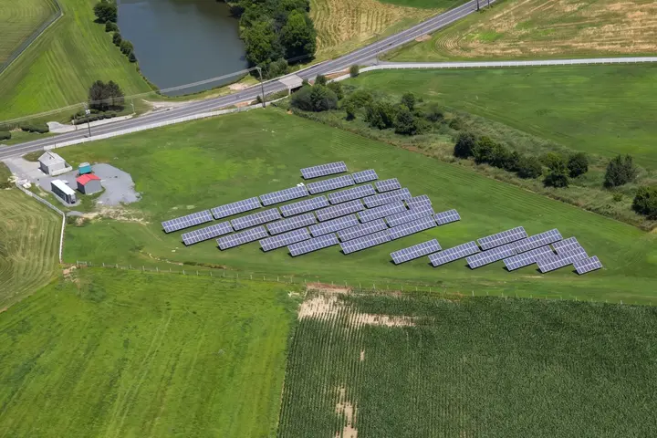 Aerial view of the Penn Township solar array: rows of tilted panels on a cut-grass field beside a country road, with sheds, a pond and trees beyond