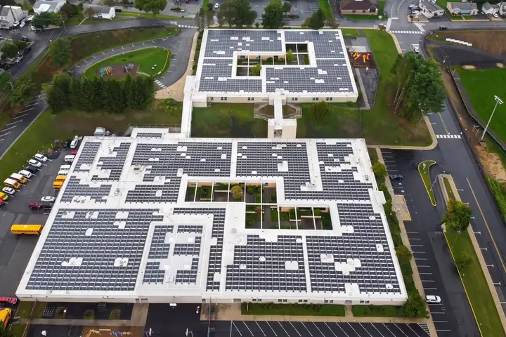 Aerial view of a school's flat white roofs almost fully covered in rows of solar panels, with parking lots, school buses and roads around it