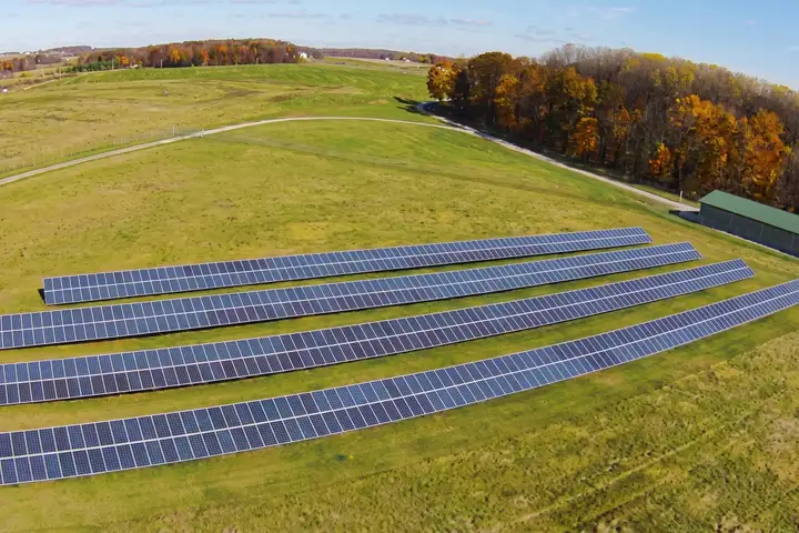 Aerial view of the York County Solid Waste Authority solar array: four long rows of tilted panels on the grassed landfill cap, with a gravel road, a green-roofed building and autumn trees beyond